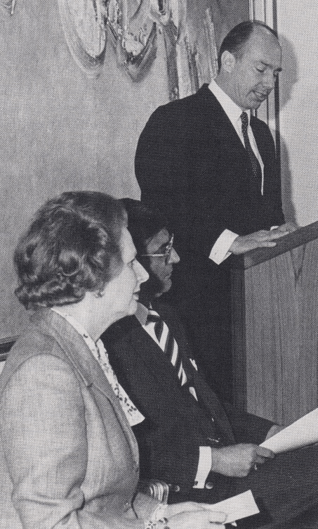 A historical black and white photograph featuring a man speaking at a podium, with two individuals seated in the foreground, including a woman wearing a suit. The scene appears to be a formal event.