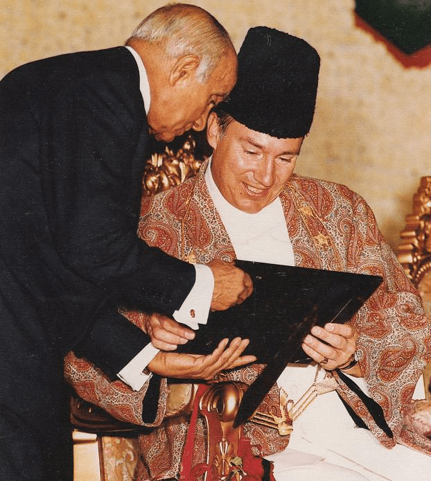 A formal meeting between two individuals, one in traditional attire and the other in a suit, sharing a document.