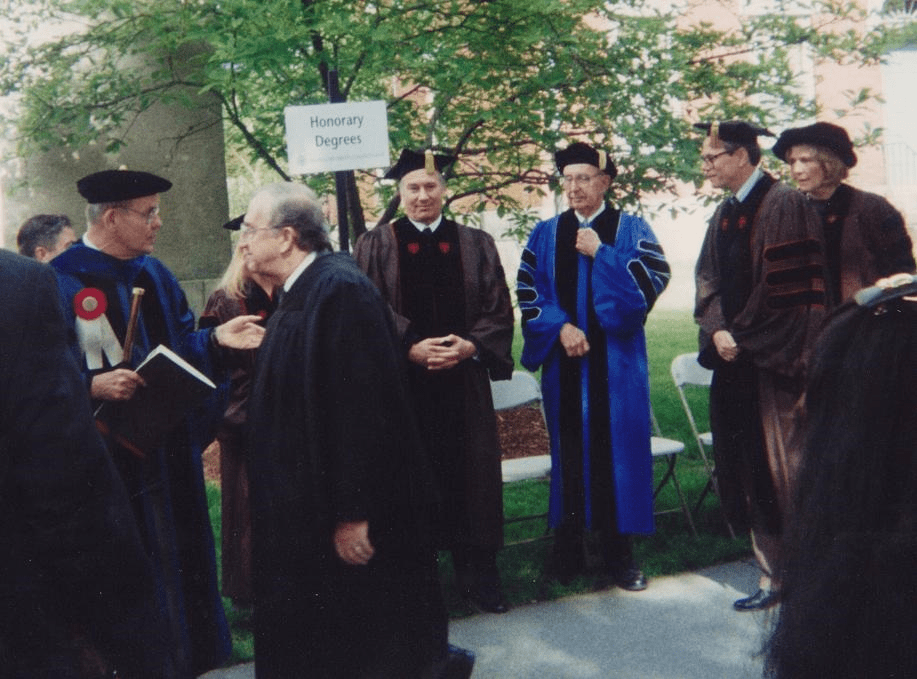 Group of individuals in academic regalia gathered outdoors, discussing near a sign labeled 'Honorary Degrees'.