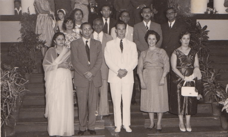 A black and white photograph depicting a group of individuals standing on a staircase, dressed in formal attire. The group includes men in suits and women in elegant dresses, posing together in a community or ceremonial setting.