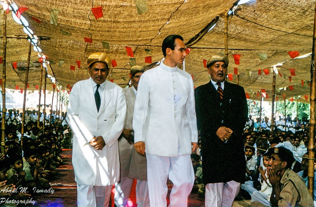 Mawlana Shah Karim walking with officials under a decorated canopy, surrounded by seated audience members during a public event.