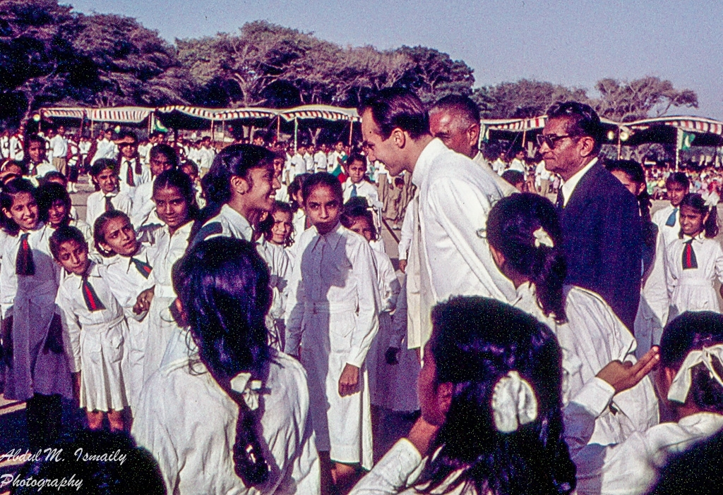 Mawlana Shah Karim (Aga Khan IV) interacting with children at a gathering, surrounded by a crowd in white attire.