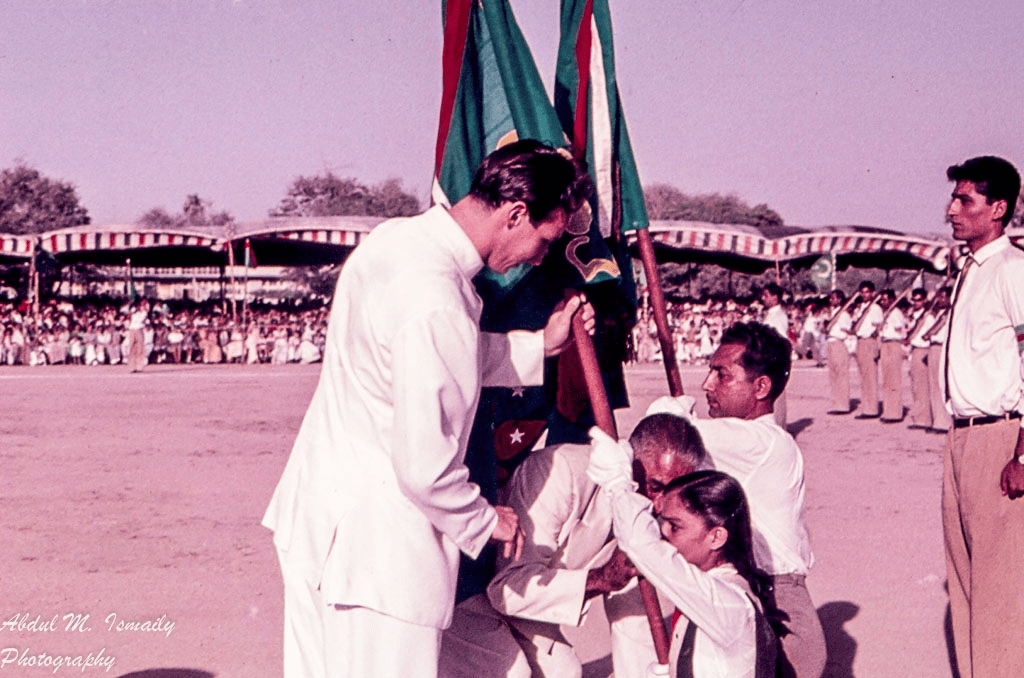 A historical photograph depicting Mawlana Shah Karim receiving a flag from young children during a ceremonial event, with a large audience in the background.
