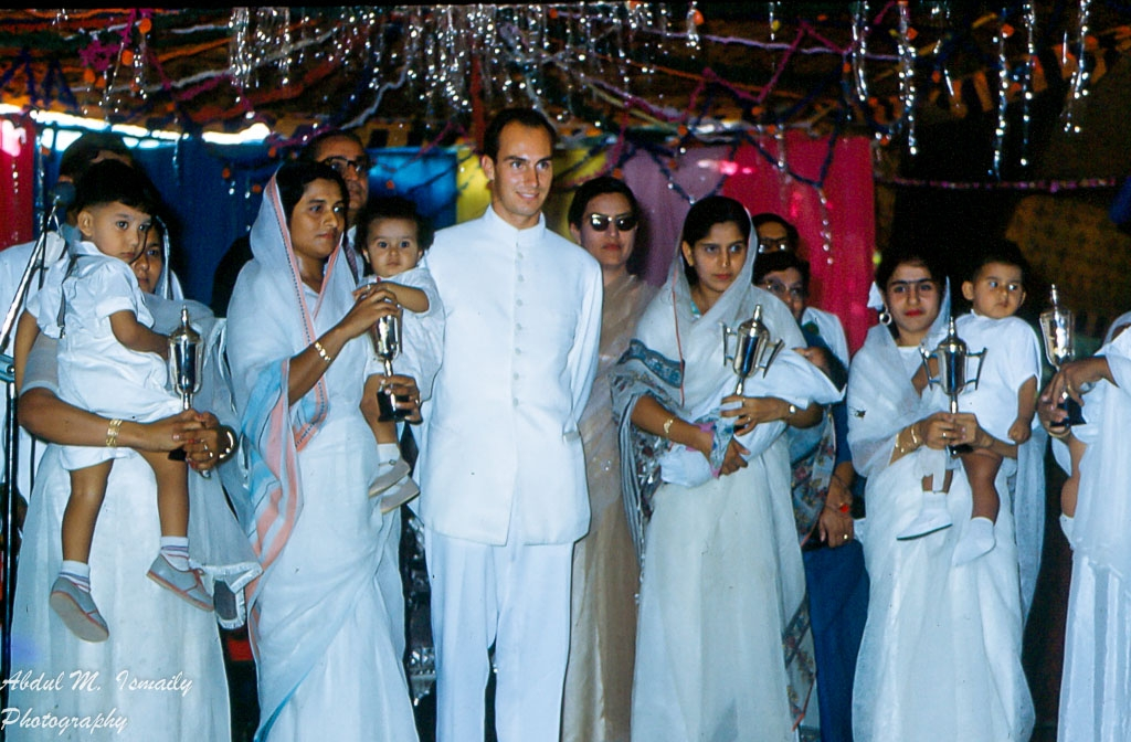 A group of women and children, dressed in traditional attire, participating in a cultural or religious event, holding candles or lanterns, with a man in white attire standing among them.