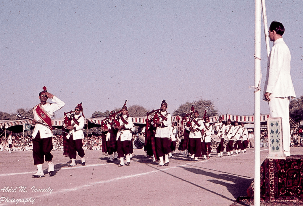 A ceremonial parade featuring a band in traditional attire, with musicians playing bagpipes and drums, moving towards a dignitary standing on a platform.