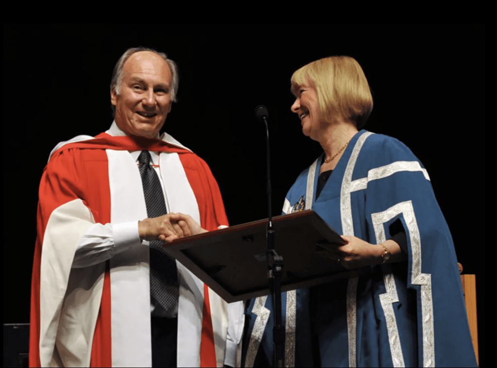 Aga Khan IV, wearing a ceremonial robe, smiles and shakes hands with a woman in academic regalia during a graduation ceremony.