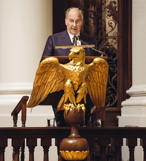 His Highness the Aga Khan speaking at a podium adorned with a golden eagle emblem.