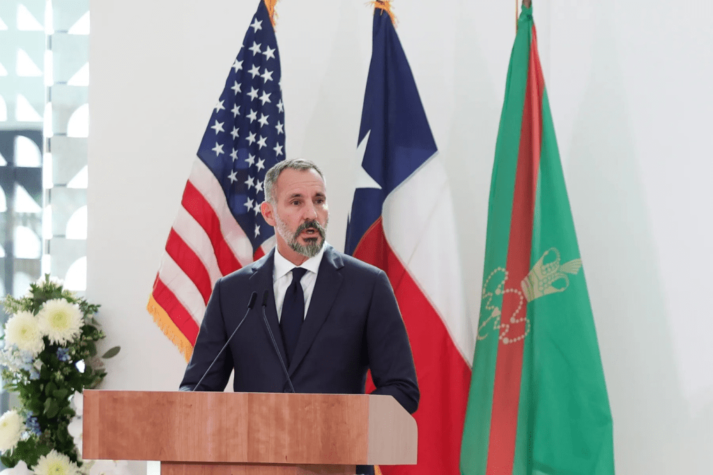A man in a suit speaking at a podium, with the flags of the United States and a green flag featuring a gold crest behind him.