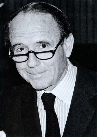 A black and white portrait of a man wearing glasses, looking directly at the camera with a slight smile.