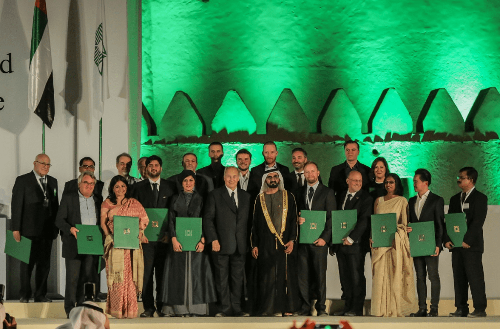 Group photo of diverse individuals holding up green folders on stage under decorative lighting.