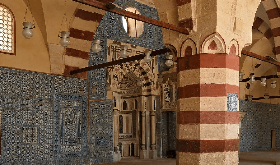 Interior view of a historical mosque featuring ornate blue tile work, striped columns, and arches.