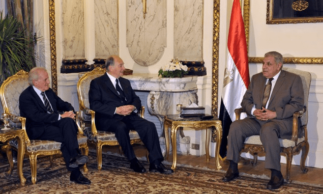 Aga Khan IV seated with two other men in a formal meeting, discussing in an elegant room adorned with Egyptian flags.