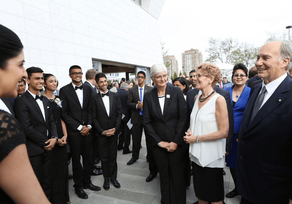 A group of formally dressed individuals smiling and interacting at an outdoor event, with a modern building in the background.