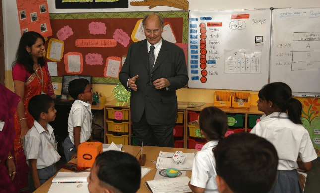 His Highness the Aga Khan interacting with school children in a classroom setting, surrounded by a few adults. Educational materials and a weekly schedule are visible on the classroom wall.