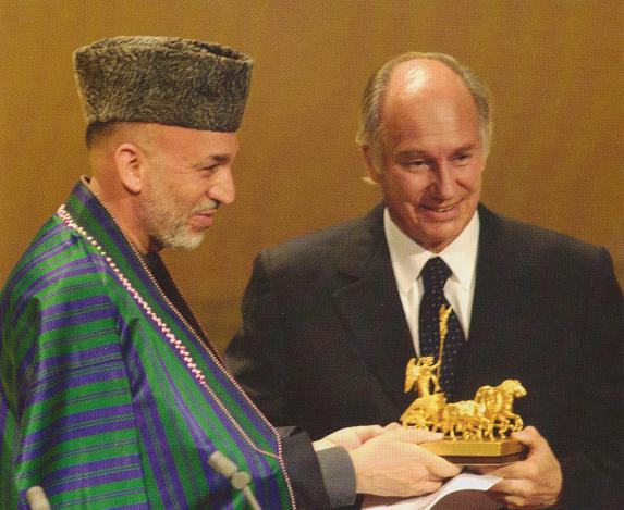 Aga Khan IV, wearing a suit, receives a golden horse sculpture from a man in traditional Afghan attire at a ceremonial event.