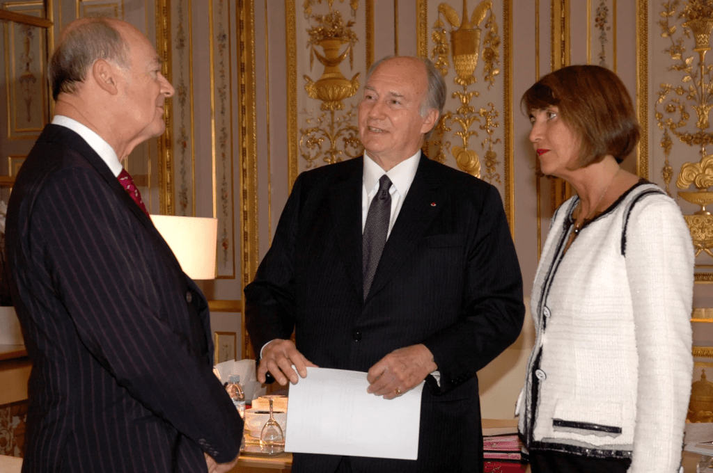 Aga Khan IV engaged in a conversation with two individuals in a formal setting, adorned with elegant decor.