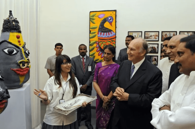 A group of people, including His Highness the Aga Khan, observing an artist presentation featuring a large decorative sculpture and paintings in a gallery setting.