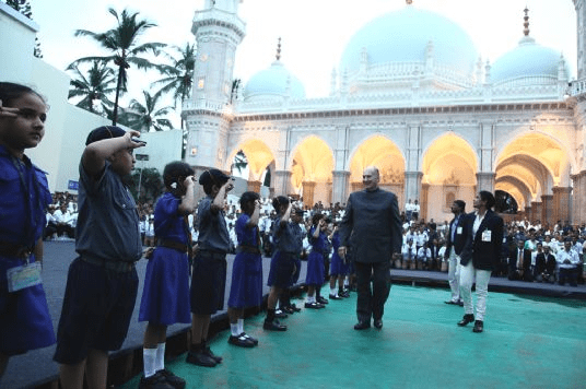 A gathering of children in uniforms saluting and greeting a distinguished figure in front of a large mosque with domes and arches.