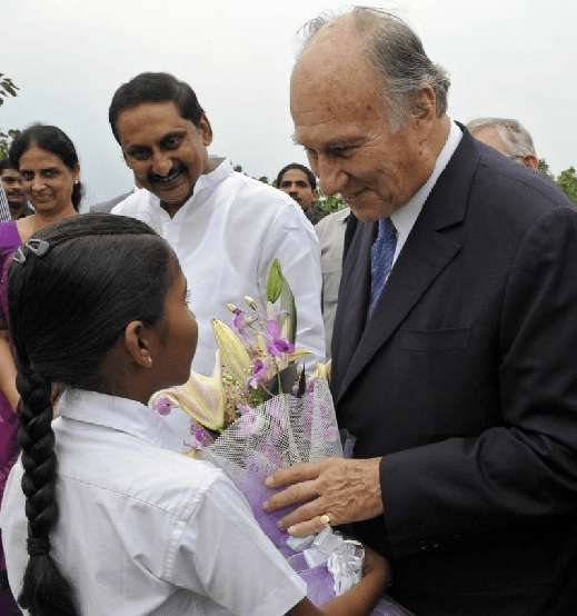 Mawlana Shah Karim, His Highness the Aga Khan, receiving flowers from a young girl during an event, with smiling individuals in the background.