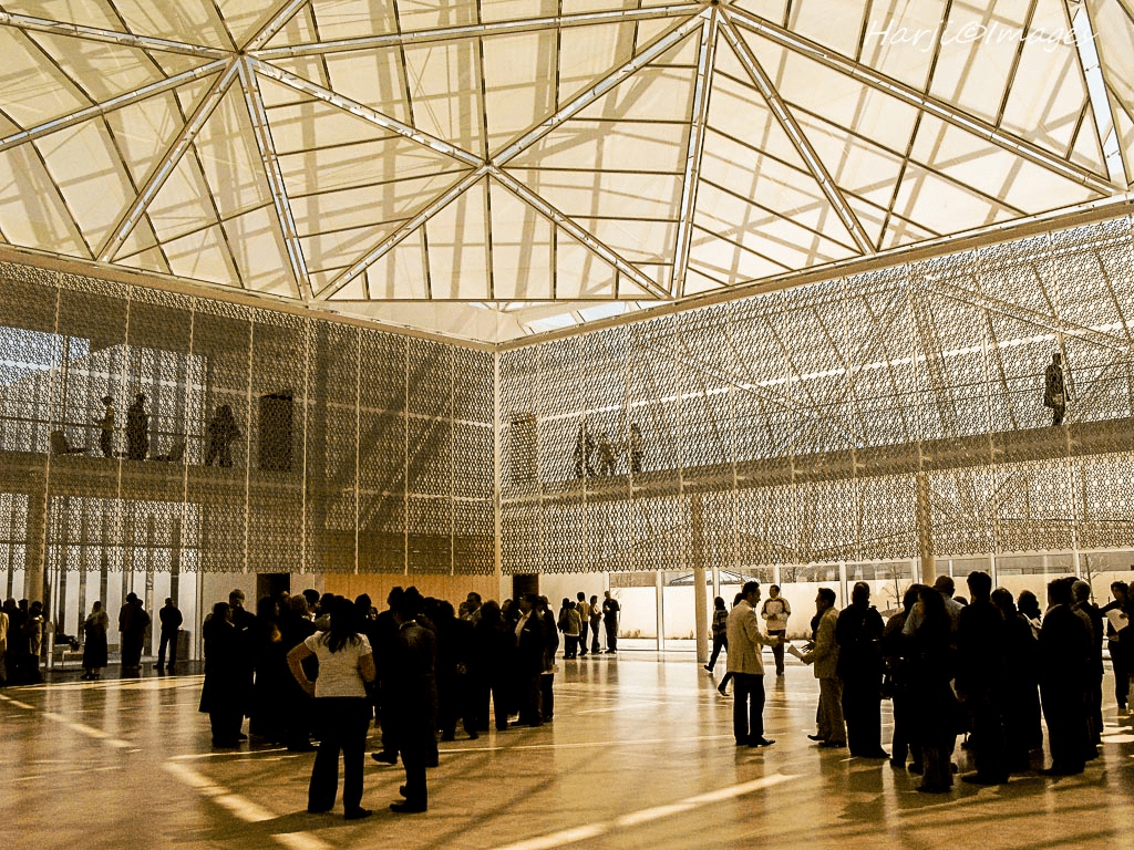 A spacious modern interior of the Ismaili Center with people gathered, featuring a high geometric ceiling and intricate lattice walls.