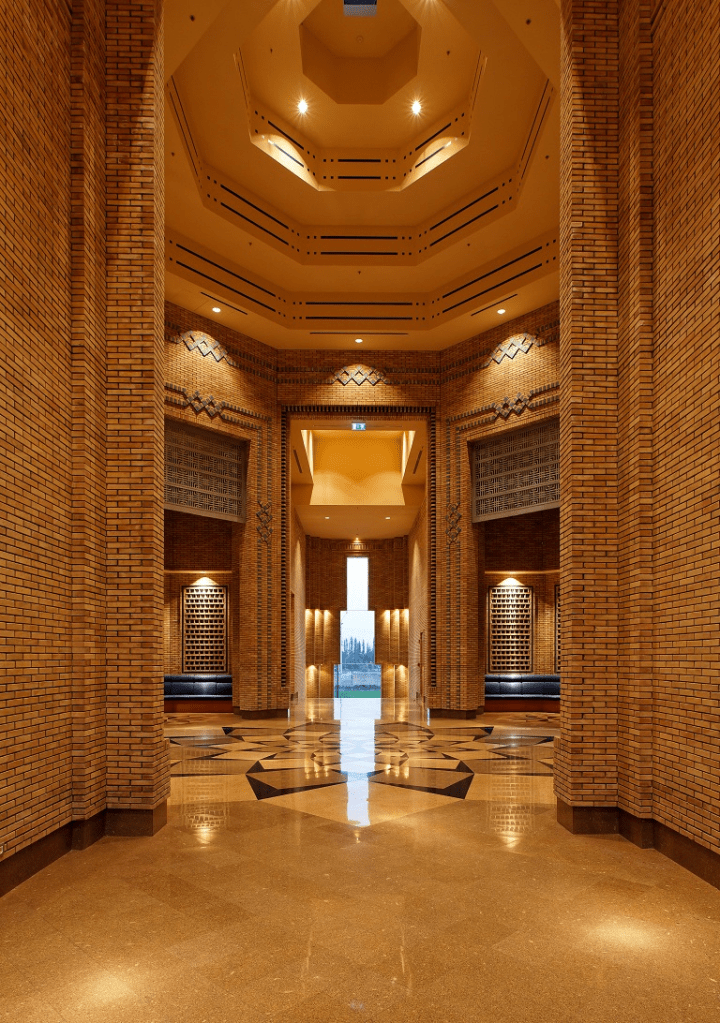 Interior view of a spacious building featuring ornate brickwork, a high ceiling with geometric patterns, and polished stone flooring, designed as a peaceful community space.