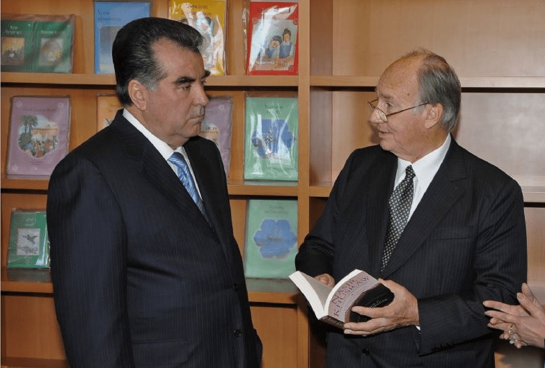 Mawlana Shah Karim, His Highness the Aga Khan IV, meeting with a dignitary while holding a book, with colorful books in the background.