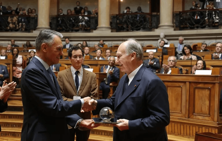 Two men shake hands in a formal setting, with one presenting a glass award to the other. An audience in the background observes the event.