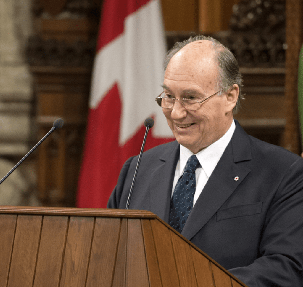 Mawlana Shah Karim, His Highness the Aga Khan, smiling during a speech at a podium in front of flags.