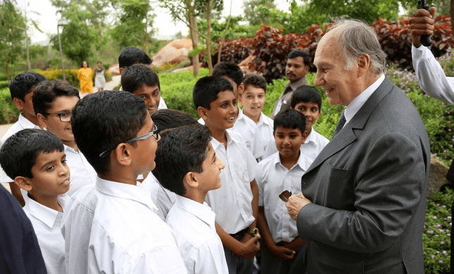 Mawlana Shah Karim, His Highness the Aga Khan IV, interacting with a group of schoolchildren in a lush green setting.