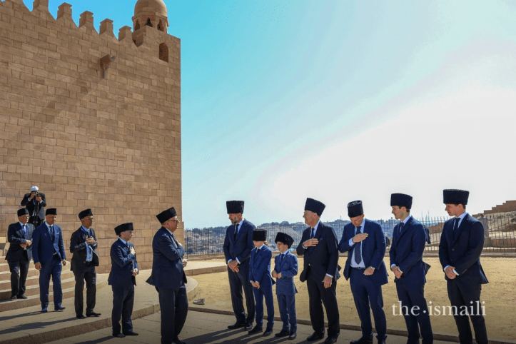 A group of people wearing formal attire and hats stand in front of a stone building, with some holding their hands over their hearts. The backdrop features a clear blue sky.
