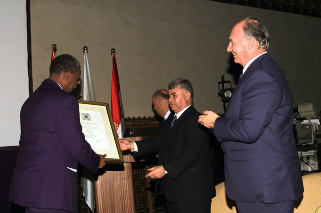 Aga Khan IV receiving an award or recognition during an event, surrounded by officials and flags.