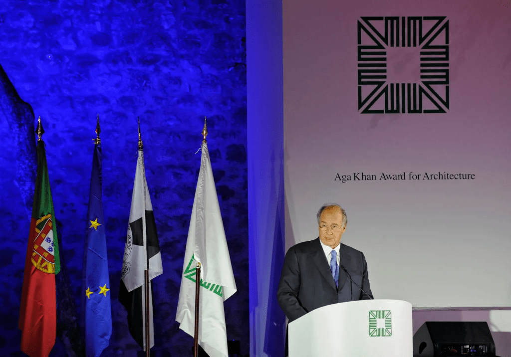Aga Khan IV speaking at the Aga Khan Award for Architecture ceremony, with flags in the background and a purple lit backdrop.