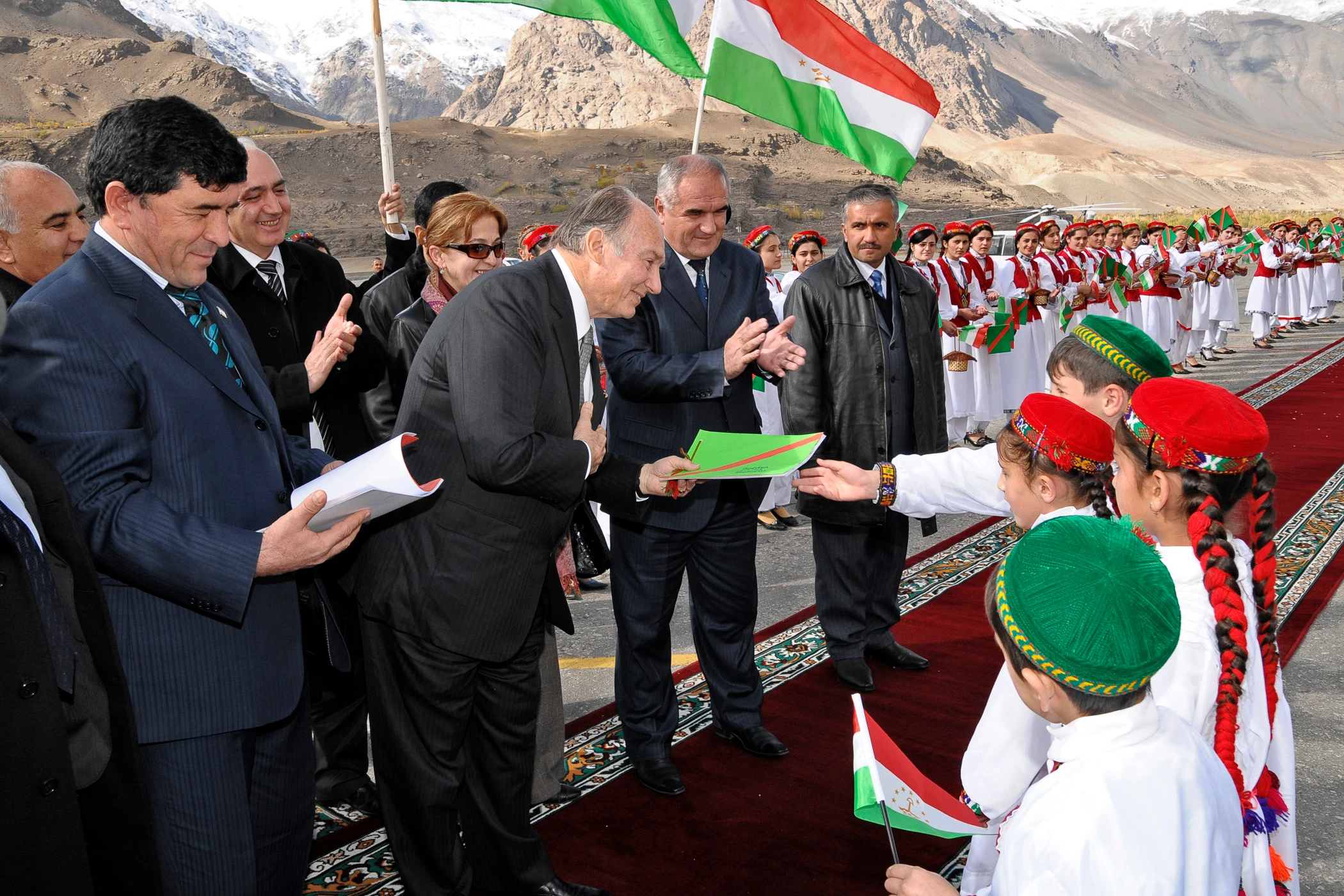 Aga Khan IV receives a warm welcome in Tajikistan from children dressed in traditional attire, holding flags, amidst a mountainous backdrop.