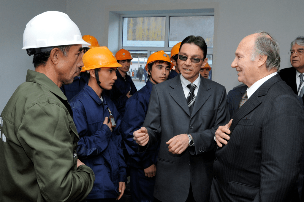 Mawlana Shah Karim, His Highness the Aga Khan IV, converses with individuals in hard hats during a visit to a construction site, surrounded by young workers and professionals.