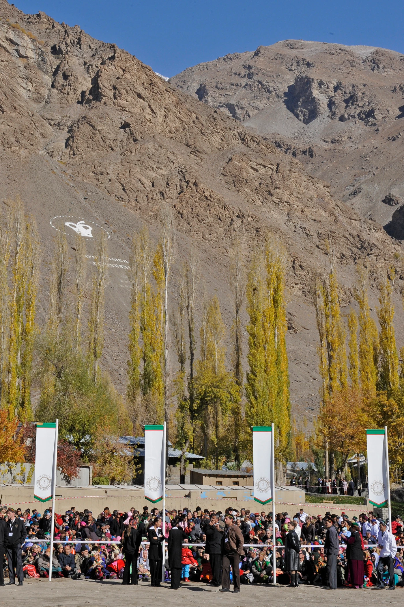 A large gathering of people attending an outdoor event in a mountainous region, with autumn trees in the foreground and a symbolic emblem painted on the rock face above.