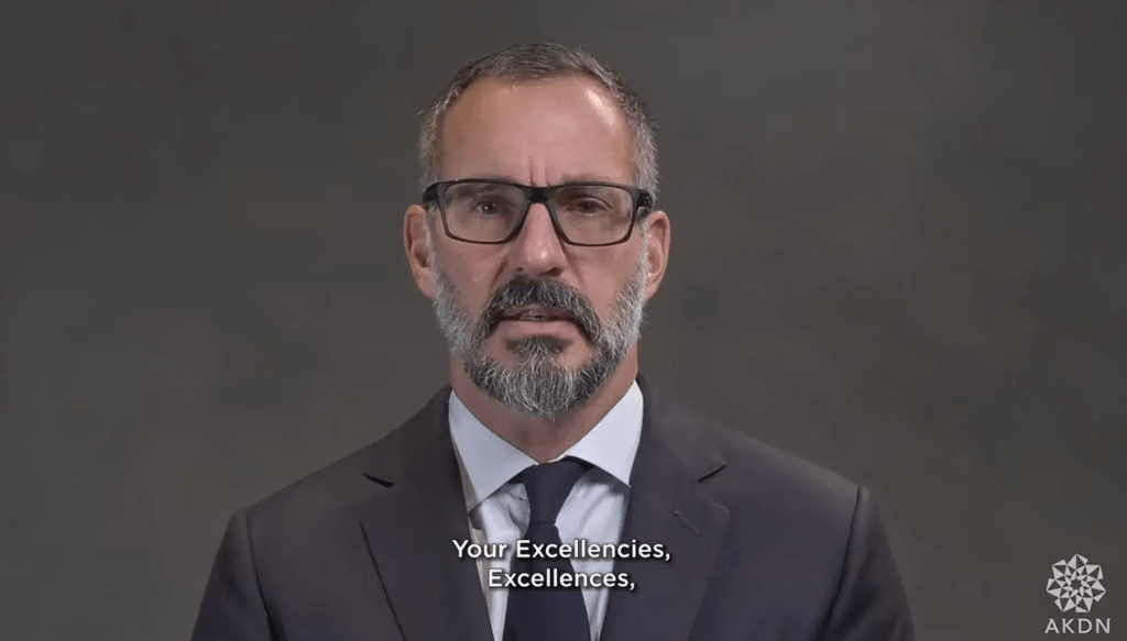 A man with glasses and a beard speaks in front of a neutral background, conveying a message.