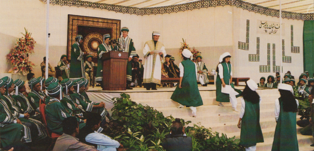 A graduation ceremony taking place on a stage with speakers and graduates wearing green robes and turbans. There are floral decorations and an audience seated in the foreground.