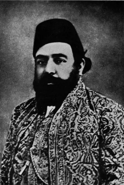 Portrait of a man wearing traditional attire with a patterned cloak and a fez hat, featuring a notable beard and serious expression.