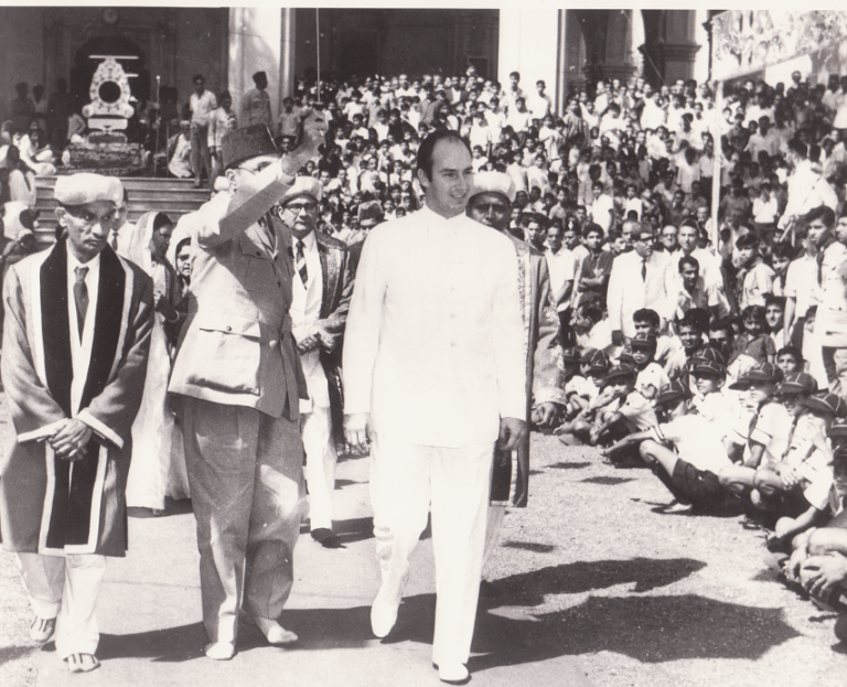 Historical image of a group of men walking in a procession, with a large crowd in the background. One man is waving, while others are dressed in traditional attire, surrounded by spectators.
