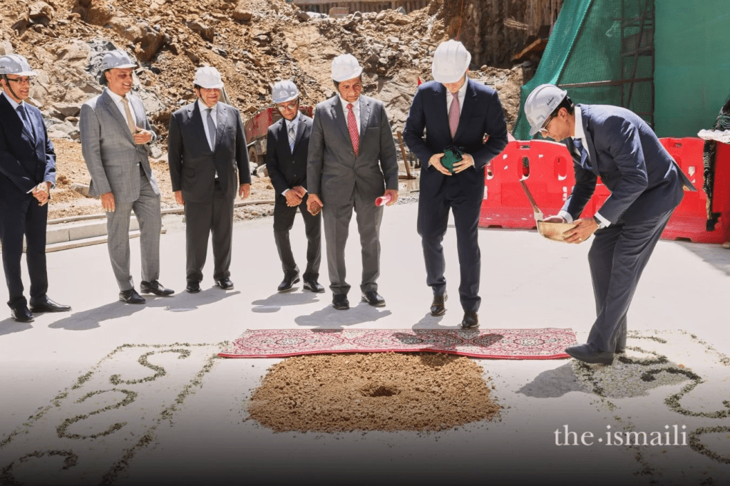 A group of six men in formal attire wearing hard hats, standing near a ceremonial ground breaking site. One man is placing a small object into a hole in the ground while others observe, and a decorative rug is laid on the ground with designs made of soil.