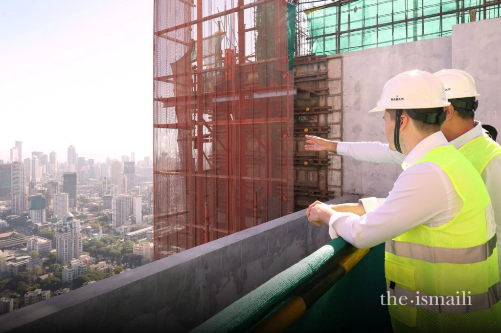 Two construction professionals in safety gear evaluate a building site from a high vantage point, overlooking a city skyline.