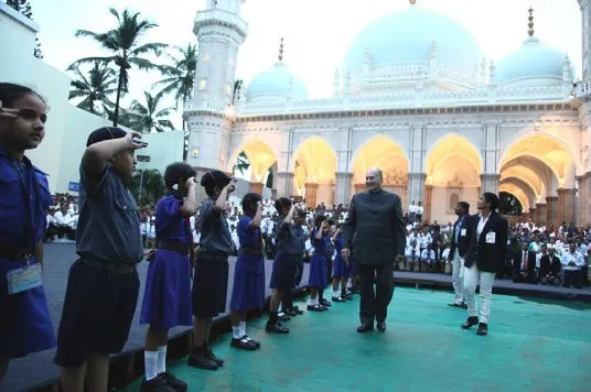 A group of children in uniforms saluting a man in formal attire outside a grand building with domes and arches, surrounded by other spectators.