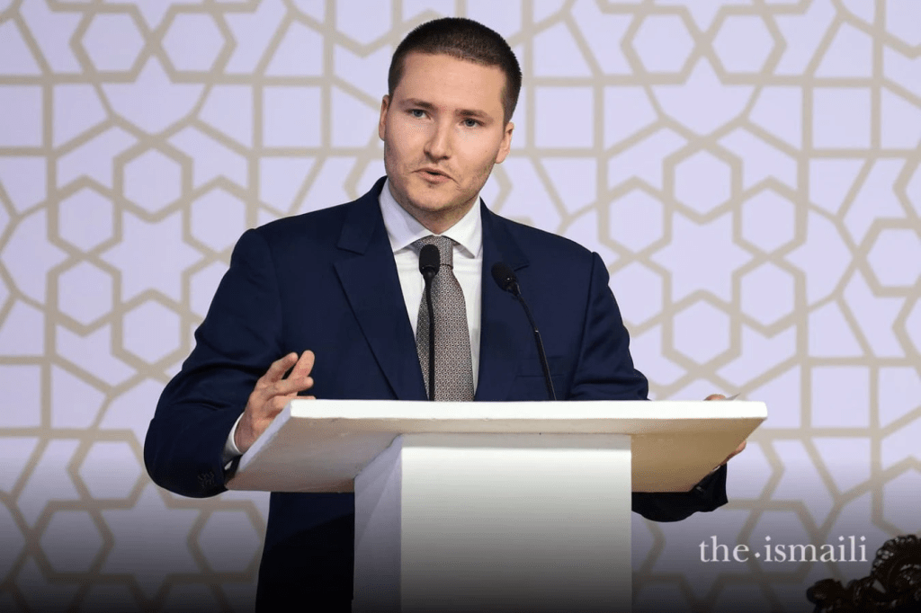 A man in a formal suit speaks at a podium against a decorative background.