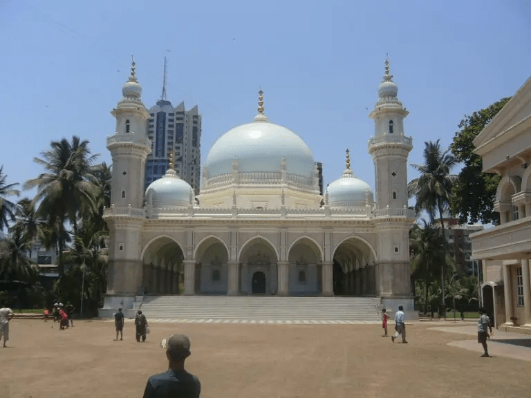 A large, ornate building with a blue dome, flanked by tall minarets, located in an urban area with palm trees and modern skyscrapers in the background.