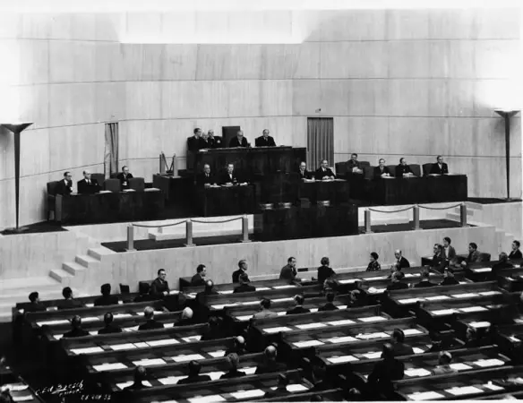 Historical black and white image of a parliamentary session, featuring a speaker at a podium, seated officials, and an audience in a legislative chamber.
